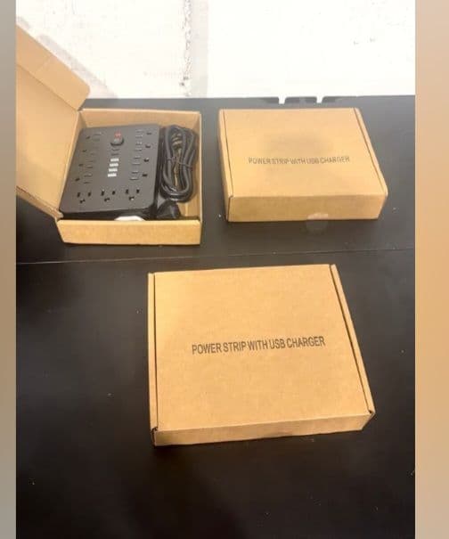 3 regletas de alimentación con cargador USB, y tipo-C ( 2 piezas negras y 1 blanca) en cajas de cartón color marrón y una unidad abierta visible en color negro, con múltiples tomas y cable de alimentación.